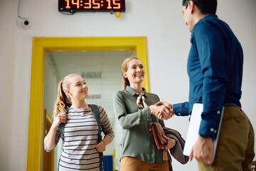Happy mother of high school student handshaking with principal in school hallway.