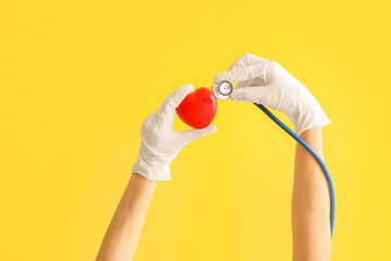 Female doctor's hands in gloves with stethoscope and heart on yellow background