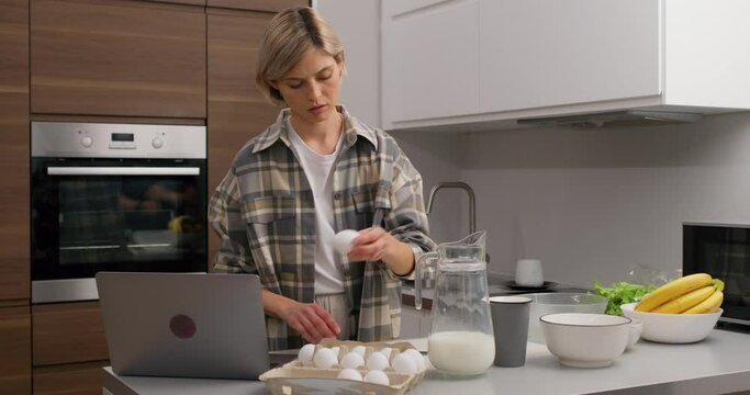 A young beautuiful fair-haired woman is cooking in a bright kitchen. She breaks egs in bowl, watching video, learning to cook, Home cooking concept