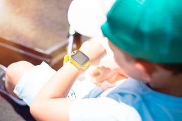 A little 7 year old boy in bright summer clothes looks at his yellow smart watch on his hand