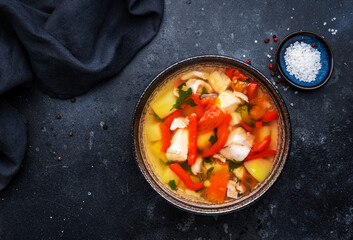 Fish soup with cod, red paprika, potatoes, tomatoes and parsley in soup bowl on black table background, top view