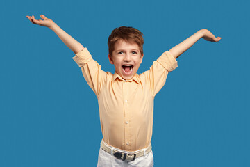 Funny little kid boy in beige shirt, posing with raised arms isolated over blue background