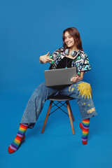 Positive lady in trendy shirt and blue jeans pointing index finger on laptop while sitting on chair, isolated over blue background. Student wearing colorful socks and having fun in studio