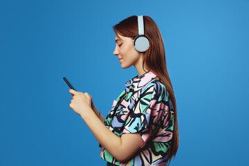 Side view of young student girl using mobile phone and looking at screen, wearing headphones while...