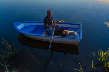 A 60-year-old Caucasian man on a blue wooden boat with oars in his hands with a poodle dog lit by the sun at sunset. - Powered by Adobe