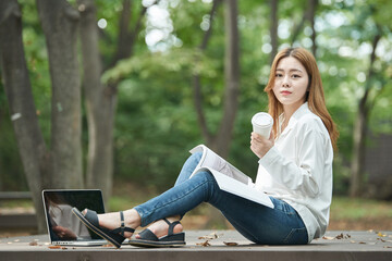 A young woman is reading a book while drinking coffee with a disposable paper cup in a forested park.