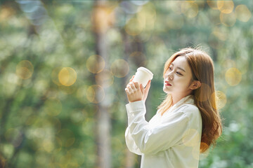 A young woman is reading a book while drinking coffee with a disposable paper cup in a forested park.