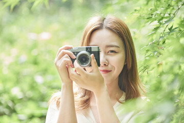 A beautiful Asian woman in a dress is taking pictures with a camera in a forested park during the summer day.
