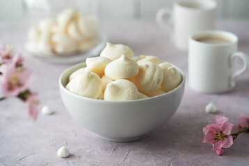 Small meringues in a white bowl over light pink background decorated with spring peach tree flowers, two cup of coffee behind - obrazy, fototapety, plakaty