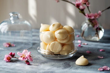 Small French meringues in a glass bowl over grey concrete background decorated with spring peach flowers, sun light behind - obrazy, fototapety, plakaty