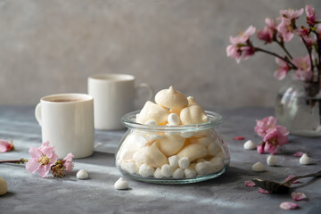 Small meringues in a glass bowl over grey concrete background decorated with spring peach flowers, cups of coffee behind - obrazy, fototapety, plakaty