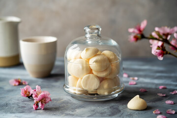 Small meringues in a glass bowl over grey concrete background decorated with spring peach flowers, cups of coffee behind - obrazy, fototapety, plakaty