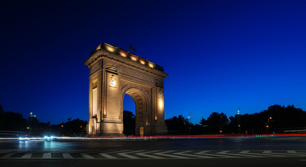 Fototapeta na wymiar Bucharest long exposure night photo. Early morning with clean blue sky at Arch of Triumph historical landmark from Romania. Traffic lights in foreground.
