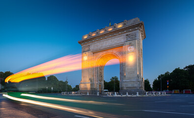 Fototapeta na wymiar Arch of Triumph in Bucharest, travel to Romania. Long exposure photo with traffic lights in the morning blue hour sky. Beautiful landscape photo of Bucharest.