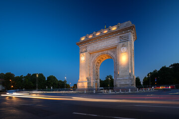 Fototapeta na wymiar Arch of Triumph in Bucharest, travel to Romania. Long exposure photo with traffic lights in the morning blue hour sky. Beautiful landscape photo of Bucharest.
