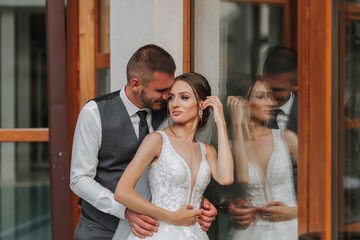 stylish bride and groom in a wedding dress with a long train and a veil are standing near a glass showcase. Front photo