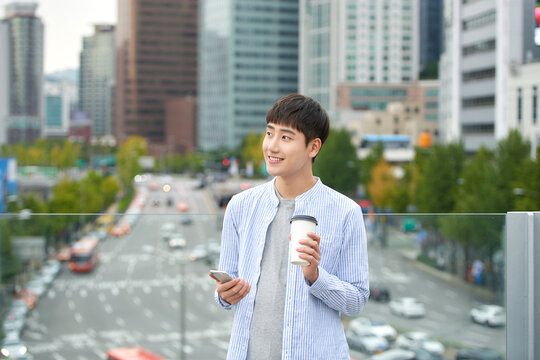 A young man is resting on a land bridge with coffee and a smartphone in the background of a busy street during the day.