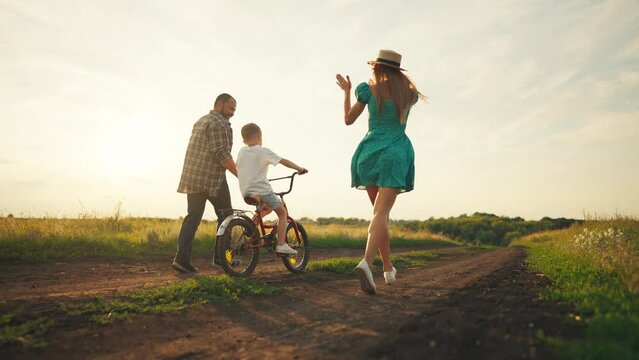 Mom dad teaching son ride bike in village on summer vacation. Parents running near child boy, mother father holding bike steering wheel supporting kid. Family weekend on nature, outdoors activity.