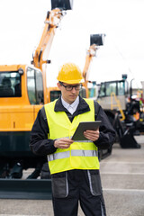 Engineer in a helmet with a digital tablet stands next to construction excavators.