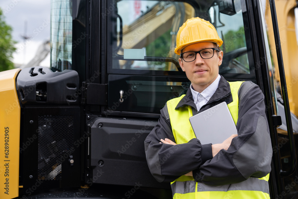 Poster Worker in a helmet with a digital tablet on the background of construction machine.