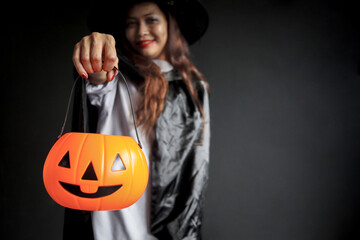 Halloween orange pumpkin in hand of beautiful attractive black long hair woman in spooky witch costume standing in dark background, fashionable female in scary dress going to celebrate Halloween party