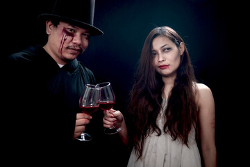 Beautiful black long hair woman white ghost spooky costume and scary black wizard man drinking red wine together in dark background, dead corpse female celebrating Halloween party with sorcerer man.
