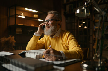 Happy elderly man talking phone while working at home office in evening