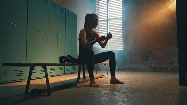 Serious woman fighter boxer wraps hands wrists with boxing bandage sits on bench in locker room, bottom view. Girl kickboxer prepares for fight. Professional sport, competition, preparation concept.
