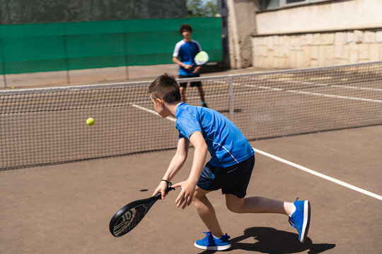 Young boy playing tennis with padel racket with unrecognizable young friend in daylight