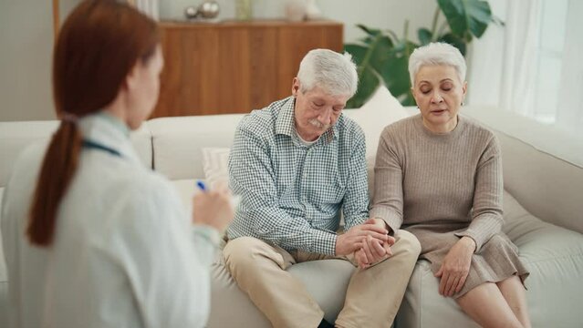 Young woman doctor nurse social worker visits senior couple. Old woman complaints about her health sits on couch with husband at home. Family support, relationship in old age and medical help concept.