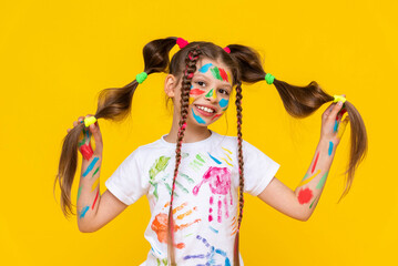 Happy little girl painted with colorful paints. A child with a stained face in paints, enjoys drawing. Art art for children and schoolchildren. Yellow isolated background.