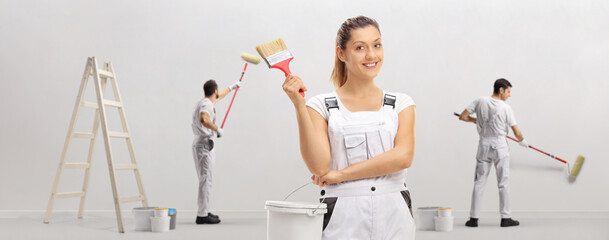 Female decorator holding a paintbrush and a color bucket and other workers painting a wall