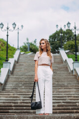 A stylish girl with fashionable makeup and hairstyle, in white trousers and a beige top and a handbag in her hands, stands against the backdrop of a large stone staircase in the park.