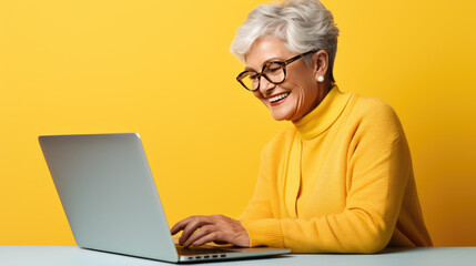 Elderly woman working on a laptop on a yellow background.