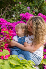 A happy mother with wavy hair holds and hugs a little blond boy with wavy hair in her arms. The baby is one year old. Against the background of flowers. Family. Mother and son