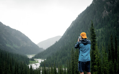 A traveler in sportswear with an orange cap. On an excursion with a view of the mountain landscape during a rainy day, fog in the background. Capturing photos on his new phone, learning photo mobile 