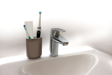 Becher mit Zahnbürste und Zahnpasta auf einem weißen Waschbecken mit Edelstahl Wasserhahn,...