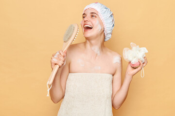 Happy funny woman wearing shower cap standing isolated over beige background being in bath posing wrapped in towel holding dry massage brush as microphone and washcloth in hand.