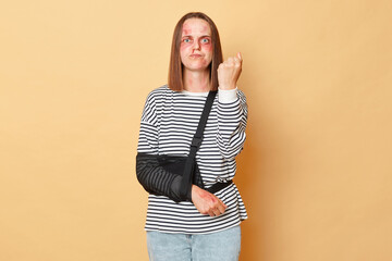 Angry sick woman with scratches and bruises on her face standing isolated over beige background showing fist after being beating revenge.