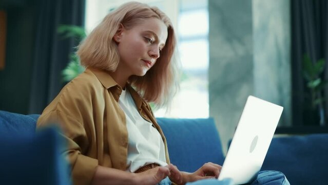 Young blonde caucasian female typing on laptop sitting on sofa
