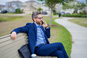 Business man talk on phone sitting on a bench in park. Man in suit call phone outside. Handsome business man using phone sitting on park bench. Business man in suit talk on phone in park.