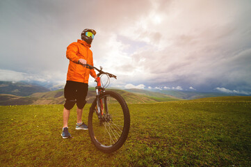 Plakat cyclist stands with a bicycle on a field at sunset in the beautiful mountains in the background. Extreme sport and enduro or downhill concept