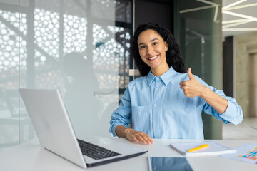 Fototapeta na wymiar Successful business woman at workplace inside office, hispanic woman smiling and looking at camera, showing thumbs up affirmatively, satisfied with achievement financial results.
