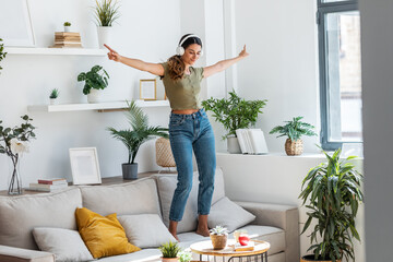 Motivated happy woman singing and dancing on couch while listening music with headphones at home.