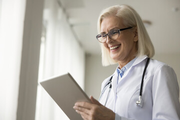 Cheerful positive senior practitioner woman holding digital device, tablet computer, talking to patient on video call, using modern Internet technology for work communication, smiling