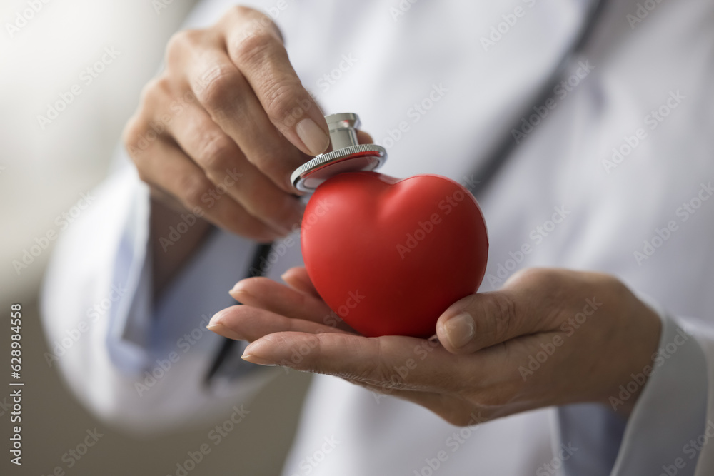 Wall mural Hands of female cardiologist applying stethoscope to red heart shaped object, offering cardiac medical checkup, heartbeat listening, early disease prevention. Close up cropped shot