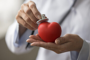 Hands of female cardiologist applying stethoscope to red heart shaped object, offering cardiac...