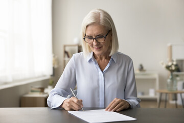 Focused successful mature old business woman in glasses signing contract, agreement at home...