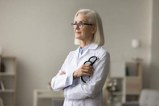 Pensive senior elder doctor woman in glasses and uniform coat looking at window away, thinking on retirement, medical occupation. Thoughtful dreamy female practitioner holding stethoscope