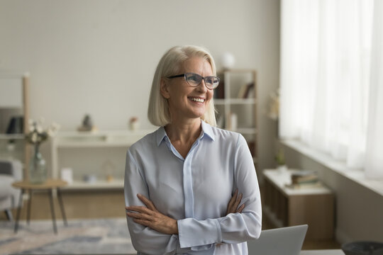 Cheerful pensive business professional woman in elegant glasses posing with folded arms indoors, looking at window away, smiling, laughing, thinking, dreaming on retirement